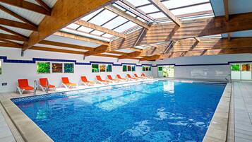 Une piscine intérieure dotée de poutres en bois et de lucarnes, entourée de chaises longues et de fenêtres offrant une vue sur la verdure.