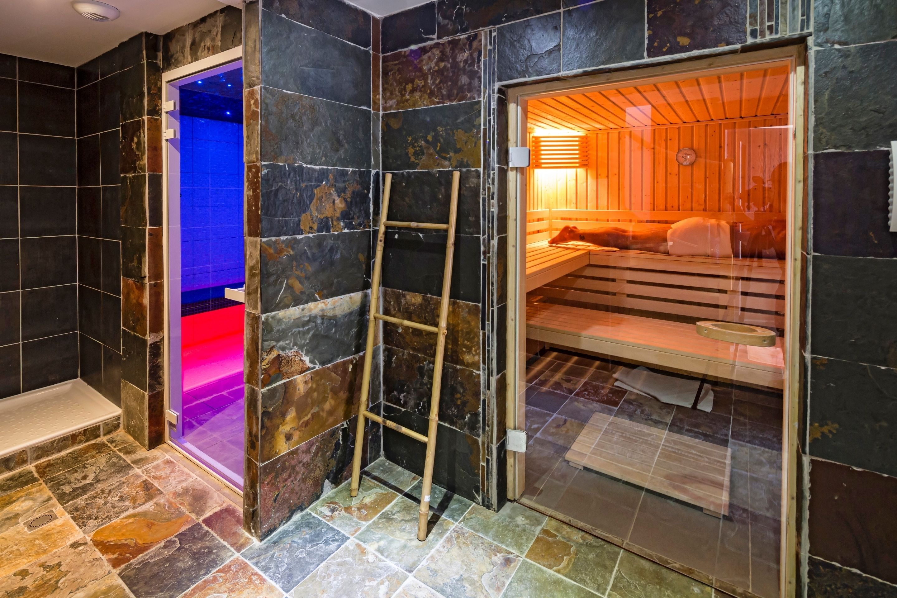 Un sauna avec un banc en bois et une échelle, un sol carrelé et une porte en verre donnant sur une salle de bain avec un espace de douche éclairé en bleu.