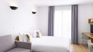 A modern hotel room with a grey sofa, a bed with white bedding, a desk with a chair, and a large window with curtains.