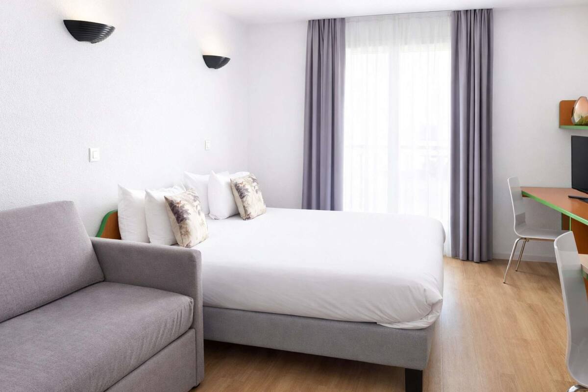 Une chambre d’hôtel moderne avec un canapé gris, un lit avec une literie blanche, un bureau avec une chaise et une grande fenêtre avec des rideaux.