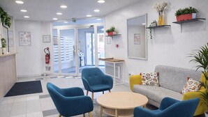 A reception area with a sofa, two blue chairs, a small table, and a coffee table. There are shelves with plants and a fire extinguisher on the wall.