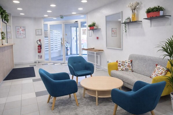Un espace d’accueil comprenant un canapé, deux fauteuils bleus, une petite table et une table basse. On y trouve également des étagères avec des plantes et un extincteur fixé au mur.