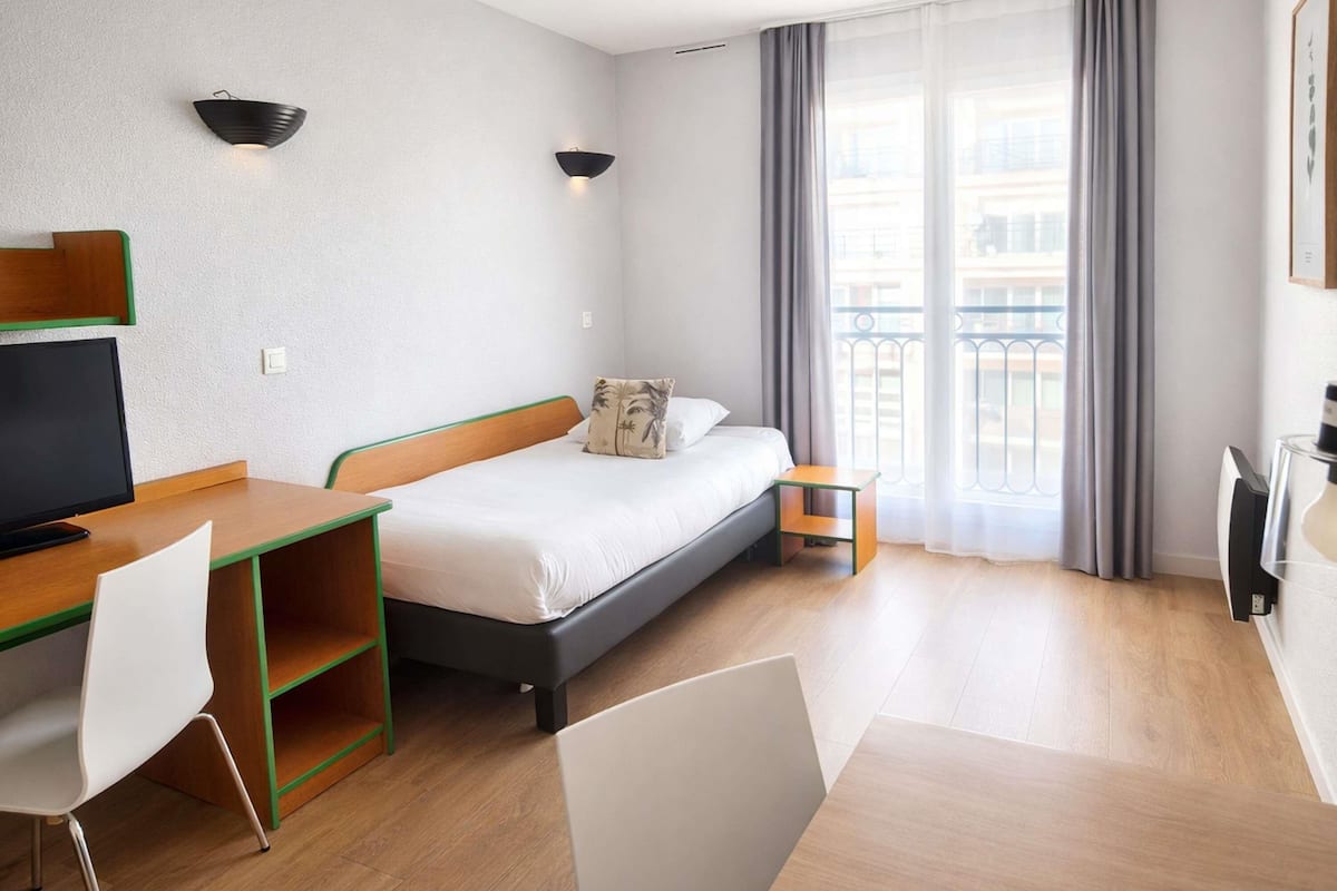 Une chambre d’hôtel avec un lit, un bureau et une chaise, une télévision et un balcon avec des rideaux.