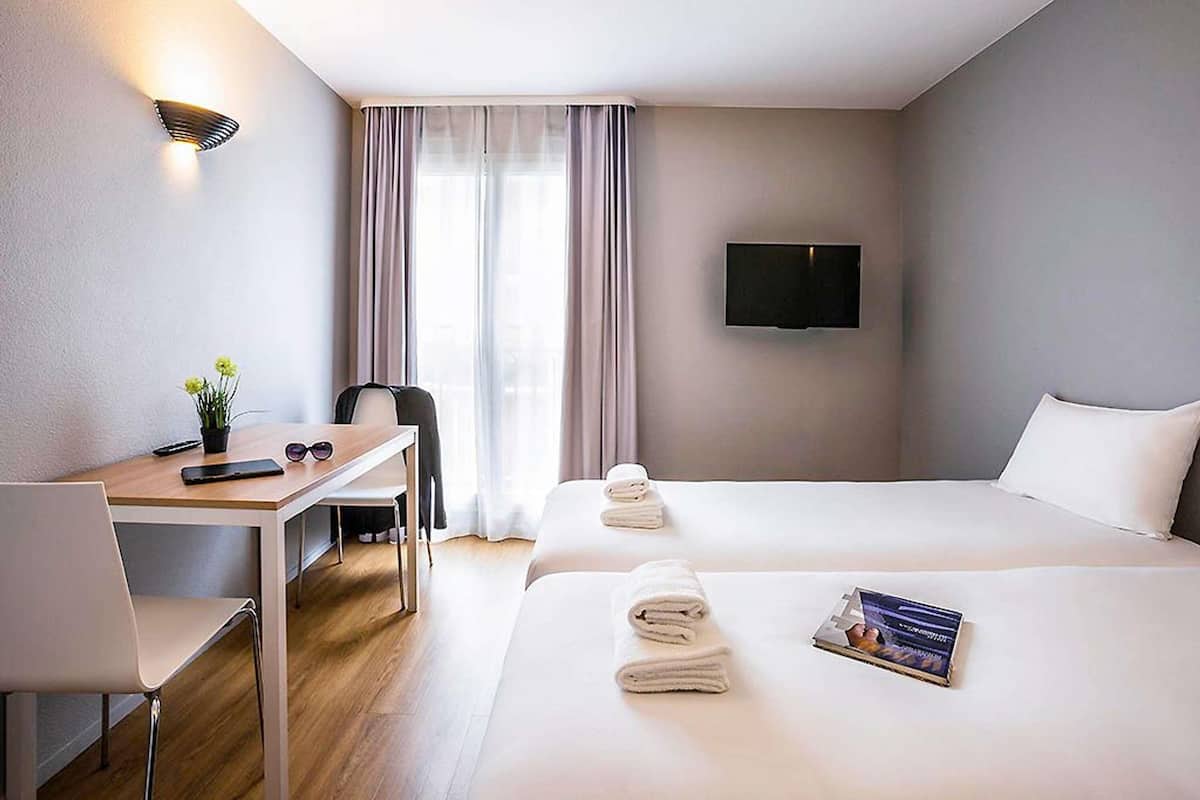 Une chambre d’hôtel équipée d’un bureau, de deux lits, d’une télévision et d’une fenêtre avec des rideaux.