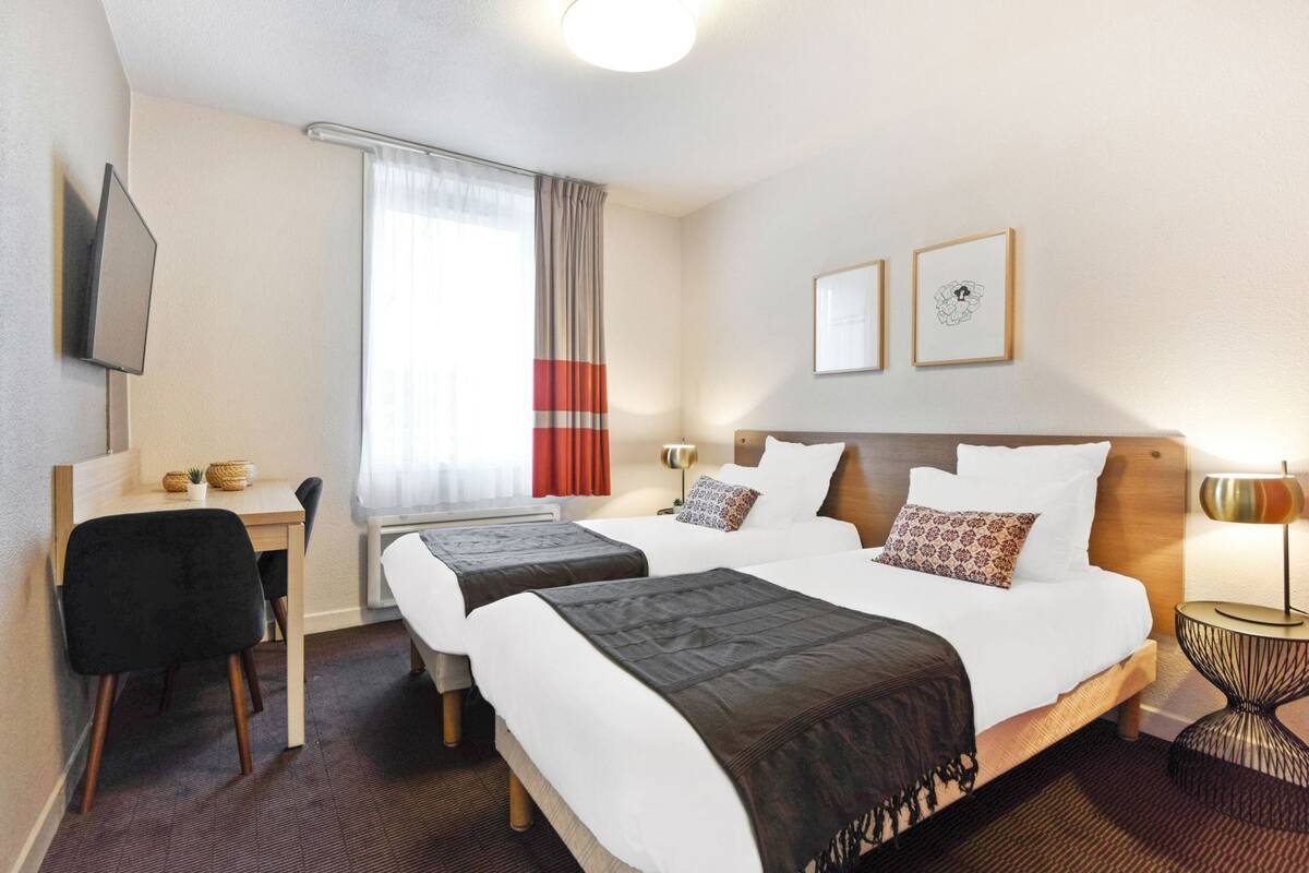 Habitación de hotel con dos camas, un escritorio, una silla, un televisor y dos cuadros en la pared.