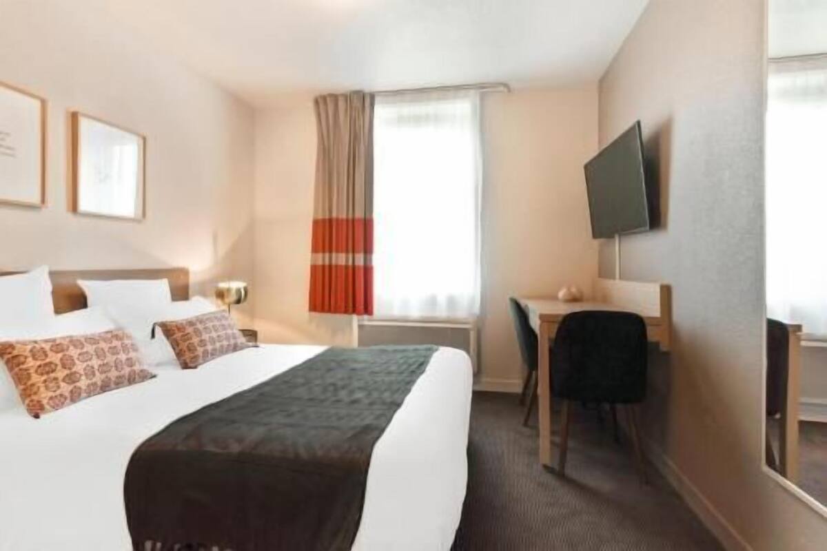 Una habitación de hotel con cama, escritorio, silla, televisor y ventana con cortinas.