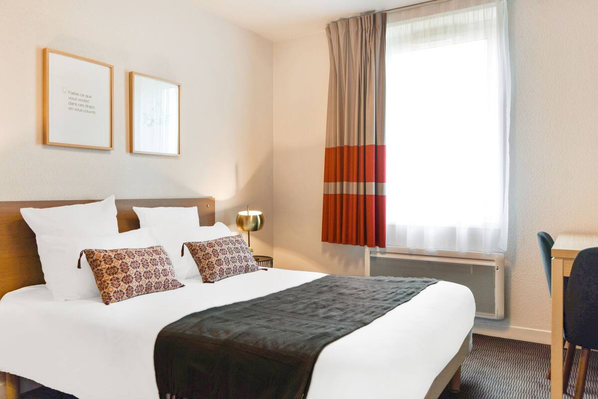 Habitación de hotel con una cama grande, dos cuadros en la pared, un escritorio con una silla y una ventana con cortinas.