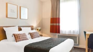 A hotel room with a large bed, two framed pictures on the wall, a desk with a chair, and a window with curtains.