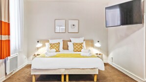 Une chambre d’hôtel avec un grand lit, deux tables de chevet avec des lampes, un téléviseur à écran plat fixé au mur et deux tableaux encadrés au-dessus du lit.