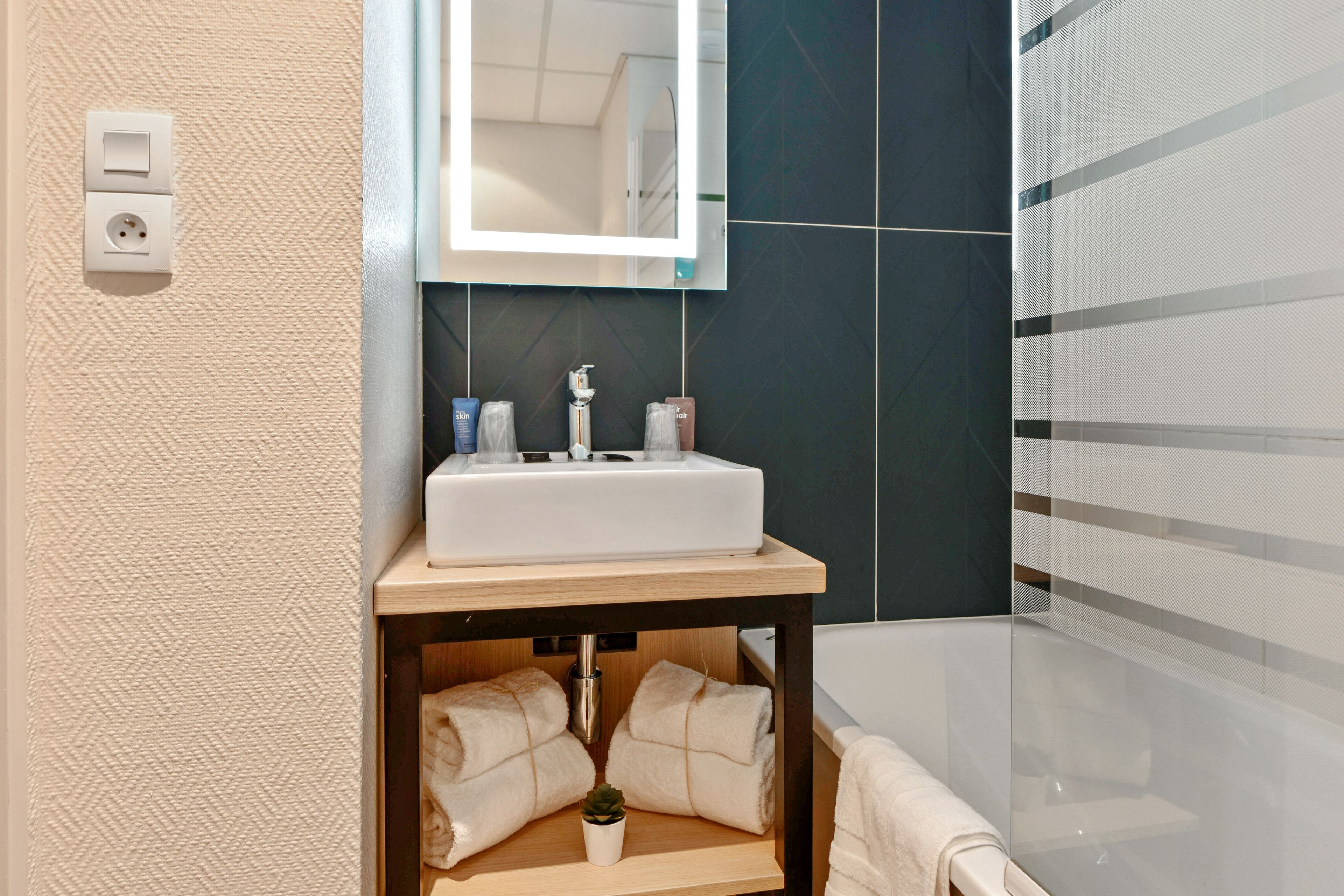 Une salle de bain avec un lavabo blanc, un miroir et un porte-serviettes. Une petite plante est posée sur l’étagère.