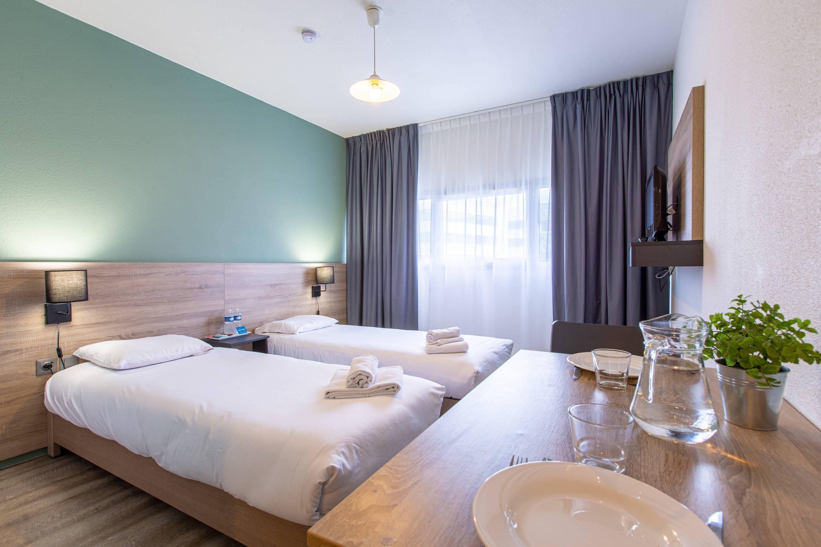 A hotel room with two beds, a wooden table with a plate and glass, a pitcher, a small plant, and a TV on the wall.