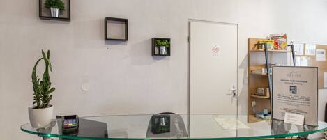 Un área de recepción con un panel de vidrio, una planta, un cuadro enmarcado, una puerta con un cartel y un tablón de anuncios.