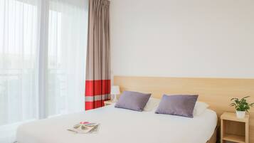 A hotel room with a large bed, a nightstand with a lamp, a small table with a plant, and a window with sheer curtains.