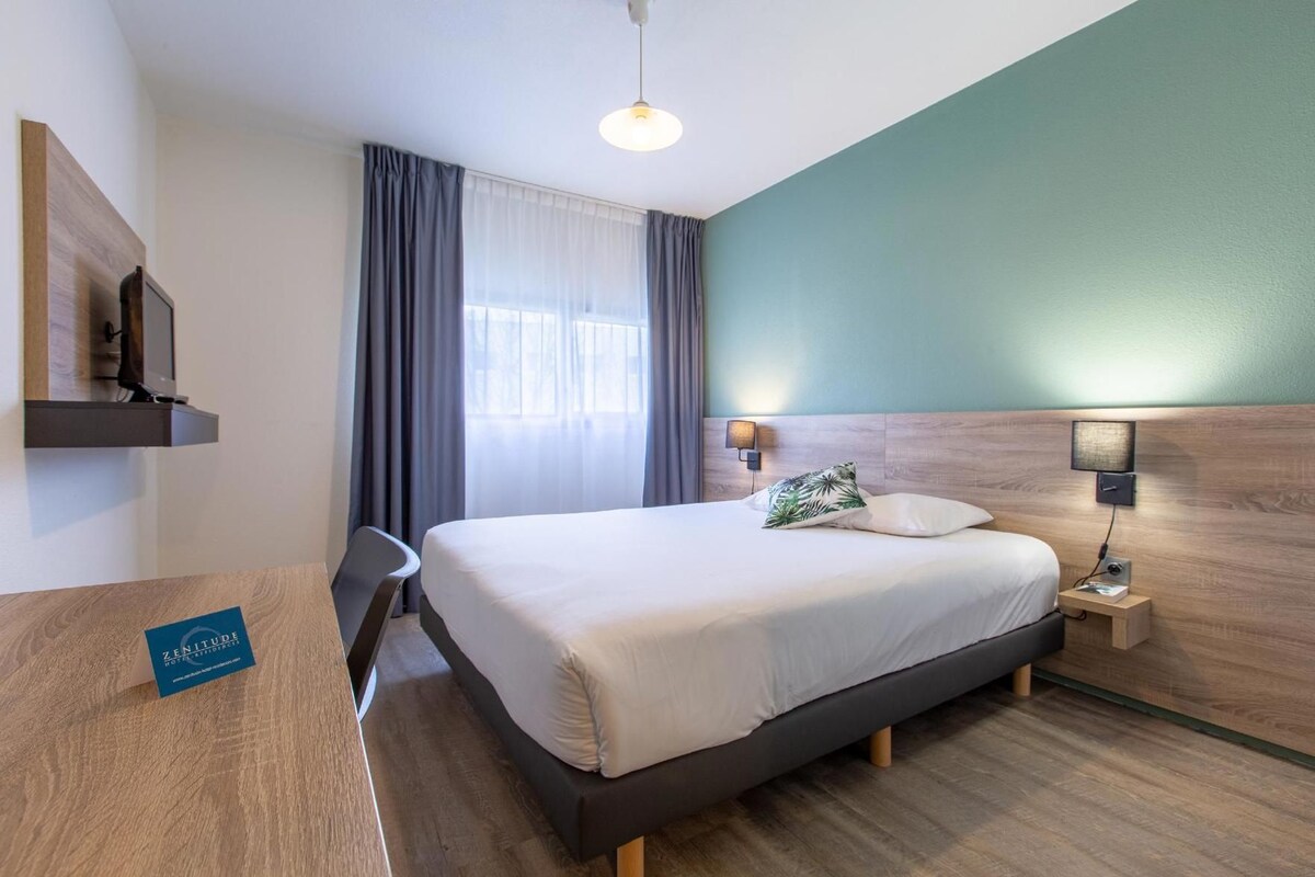 A hotel room with a bed, bedside tables, a desk, a chair, a TV, and a window with curtains.
