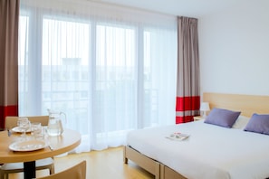 A hotel room with a bed, a table with a pitcher and glasses, and a view through sheer curtains. - Comfort Aparthotel Le Mans (Le Mans)