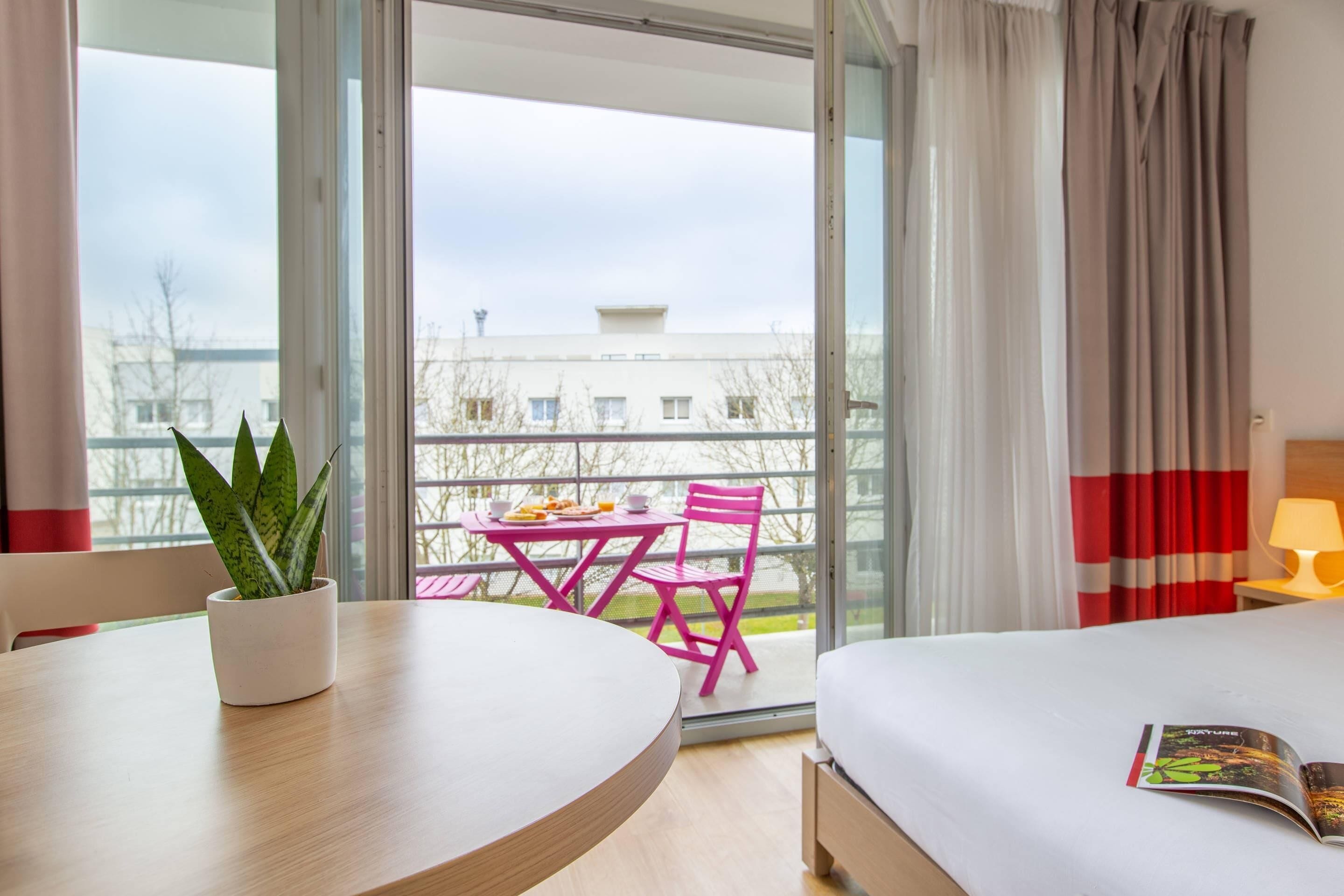 A hotel room with a bed, a table with a potted plant, a balcony with a table and chairs, and a view of buildings outside.