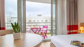 A hotel room with a bed, a table with a potted plant, a balcony with a table and chairs, and a view of buildings outside.