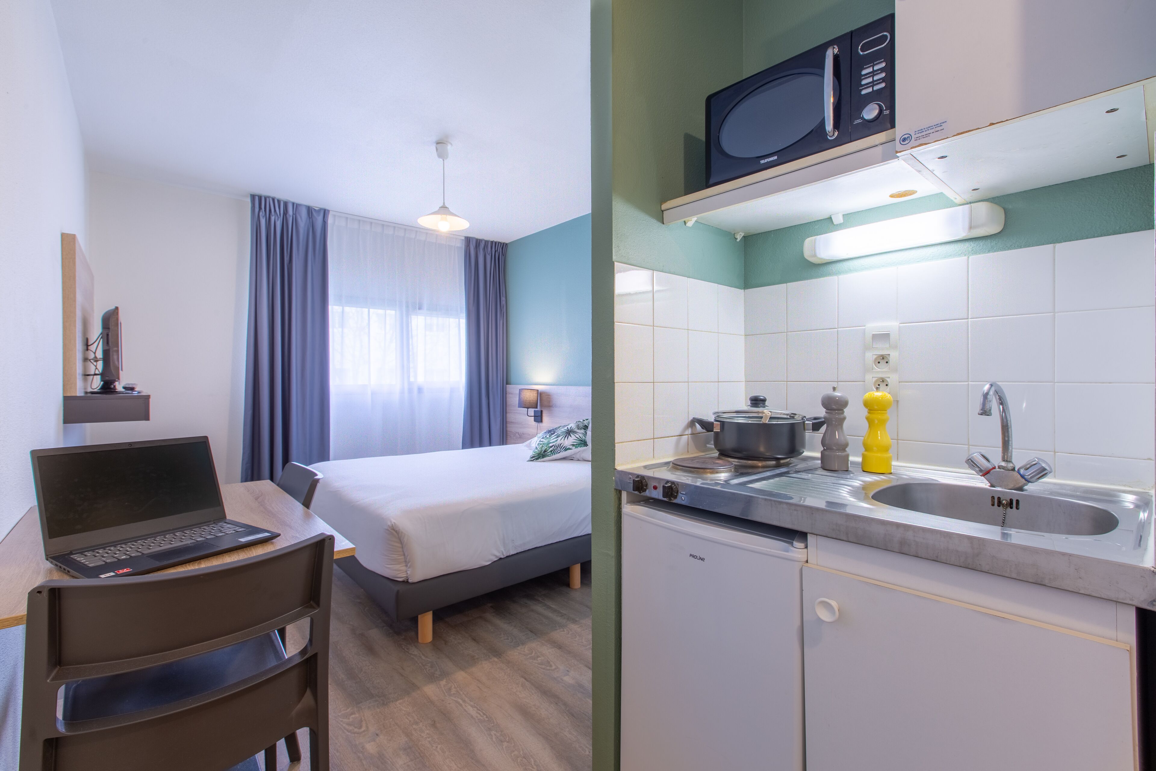 A hotel room with a bed, a desk with a laptop, a chair, a window with curtains, and a kitchenette with a microwave, stove, and sink.