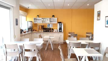 A modern cafe with white tables and chairs, a kitchen area with appliances, and a yellow accent wall.