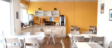 Um café moderno com mesas e cadeiras brancas, área de cozinha com eletrodomésticos e uma parede amarela de destaque.