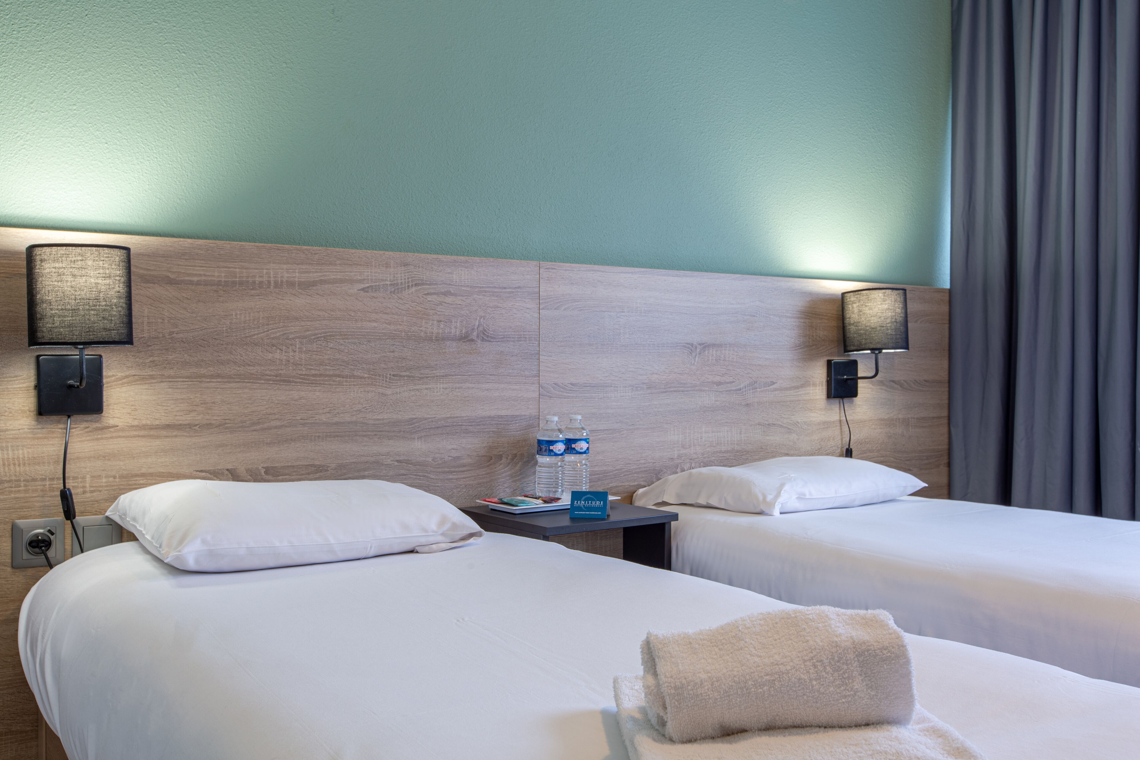 Une chambre d’hôtel avec deux lits, une table de chevet avec une lampe, une bouteille d’eau et un panneau.