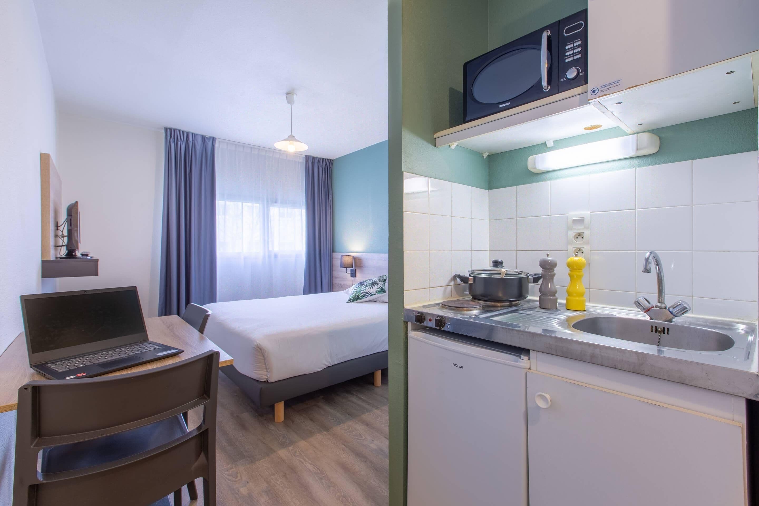 A hotel room with a bed, a desk with a laptop, a chair, a window with curtains, and a kitchenette with a microwave, stove, and sink.