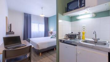 A hotel room with a bed, a desk with a laptop, a chair, a window with curtains, and a kitchenette with a microwave, stove, and sink.