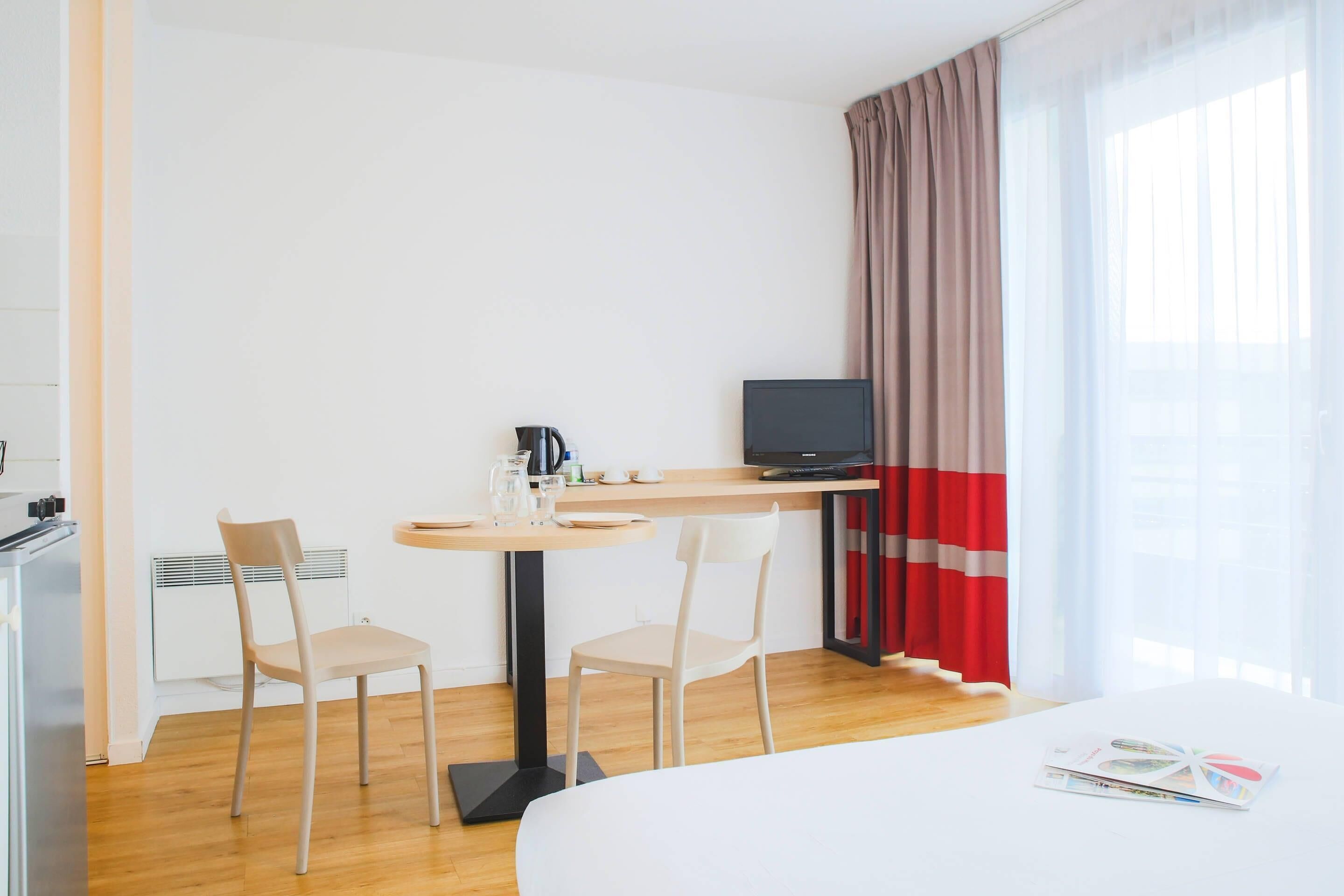 A hotel room with a bed, a desk with a computer, two chairs, a coffee maker, and a window with curtains.
