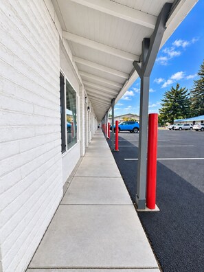 A covered walkway with a white building on one side and a parking lot with cars on the other.