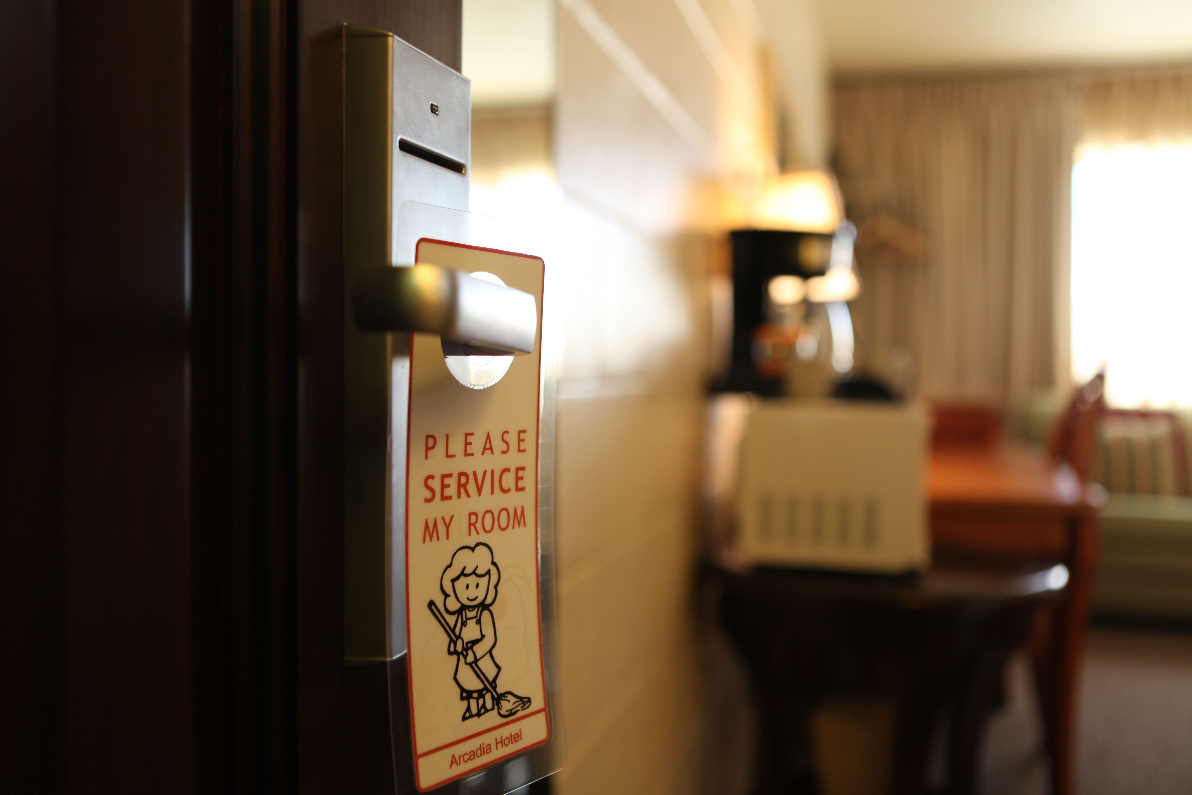 Eine Hotelzimmertür mit einem Schild, das zur Zimmerreinigung auffordert, sowie eine Lampe, ein Tisch und ein Stuhl im Hintergrund.