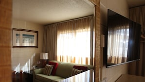 A living room with a sofa, a television mounted on the wall, and a window with curtains. - Arcadia Hotel (Gering)