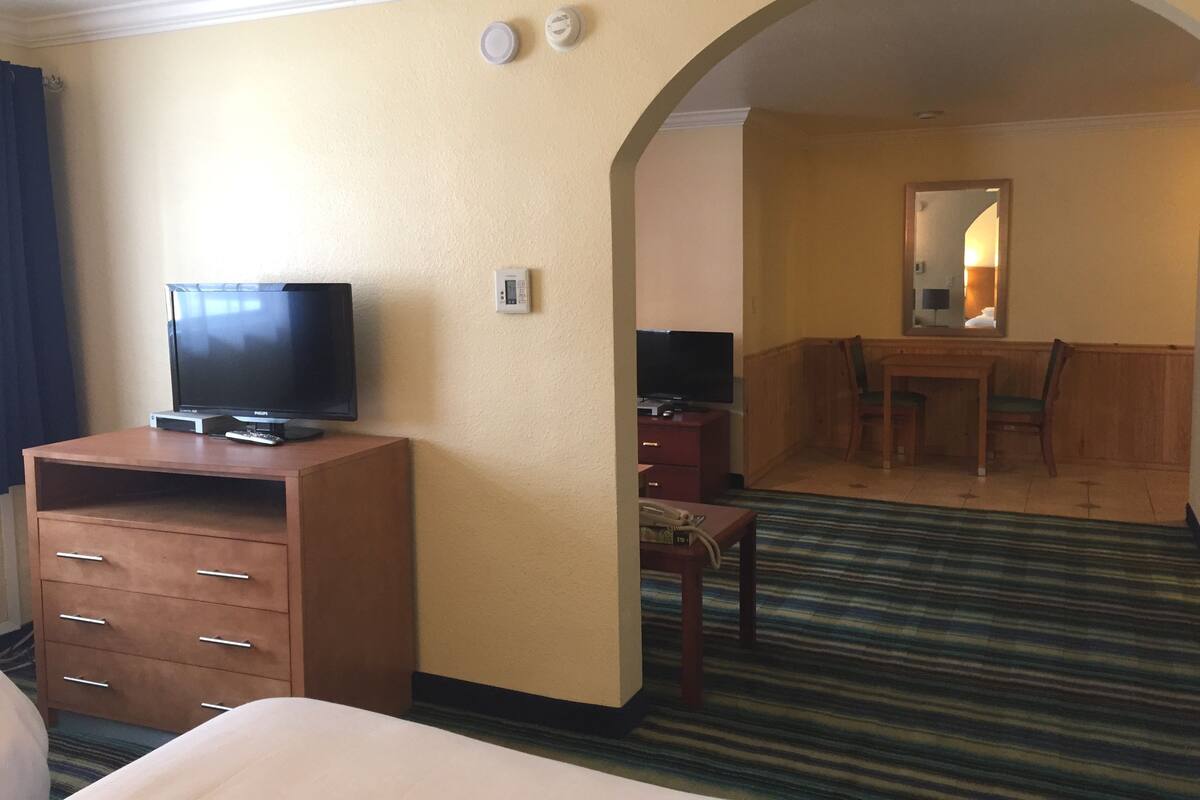 A hotel room with a bed, a television on a wooden stand, a desk with a chair, and a mirror on the wall.