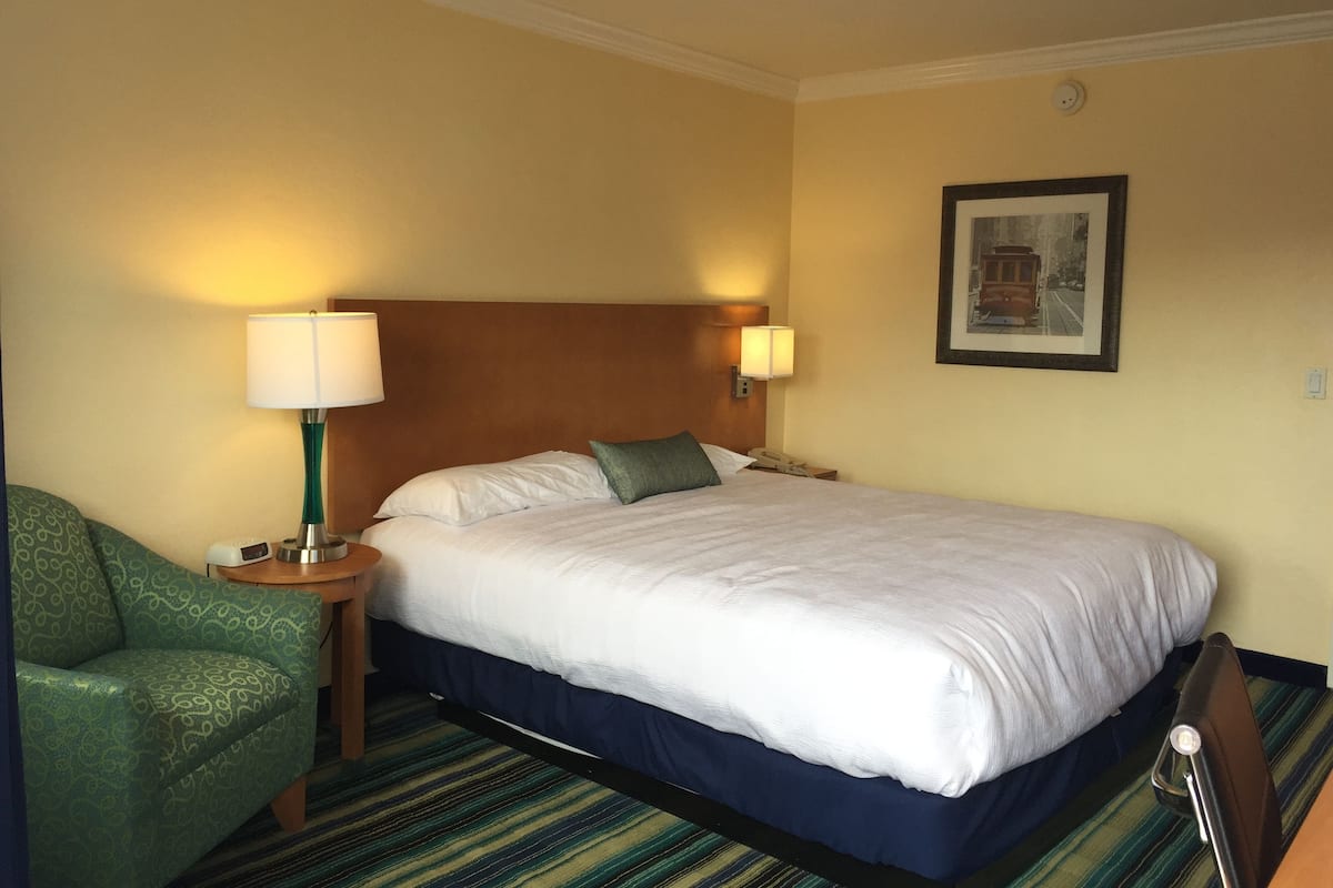 A hotel room with a large bed, two bedside lamps, a green armchair, and a framed picture on the wall.