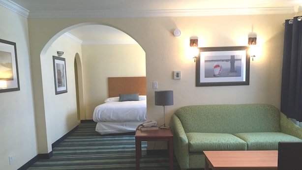A hotel room with a bed, a sofa, a small table, and a lamp. There is a framed picture on the wall and a carpeted floor.