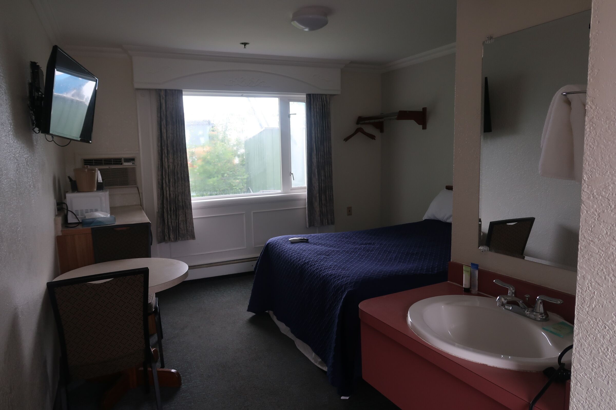A hotel room with a bed, a desk, a chair, a sink, a mirror, a television, and a window.