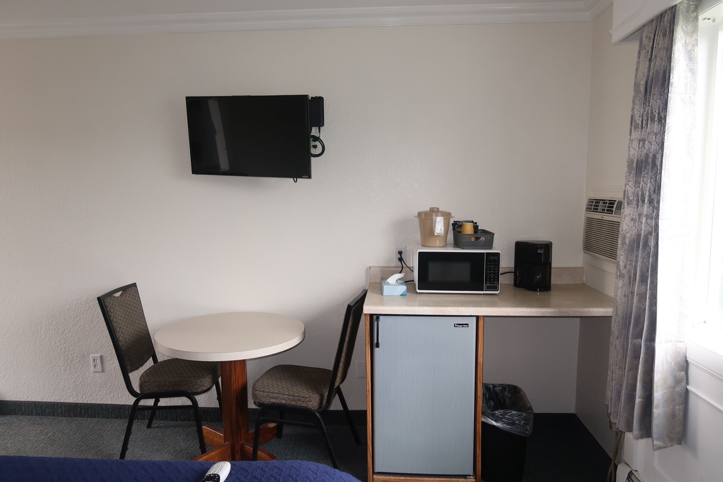 A compact hotel room with a single bed, a small desk with a microwave, a coffee maker, and a TV mounted on the wall.