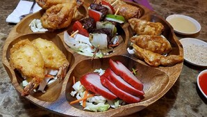 Una bandeja de madera con diversos alimentos fritos, entre ellos alitas de pollo, pescado y verduras.