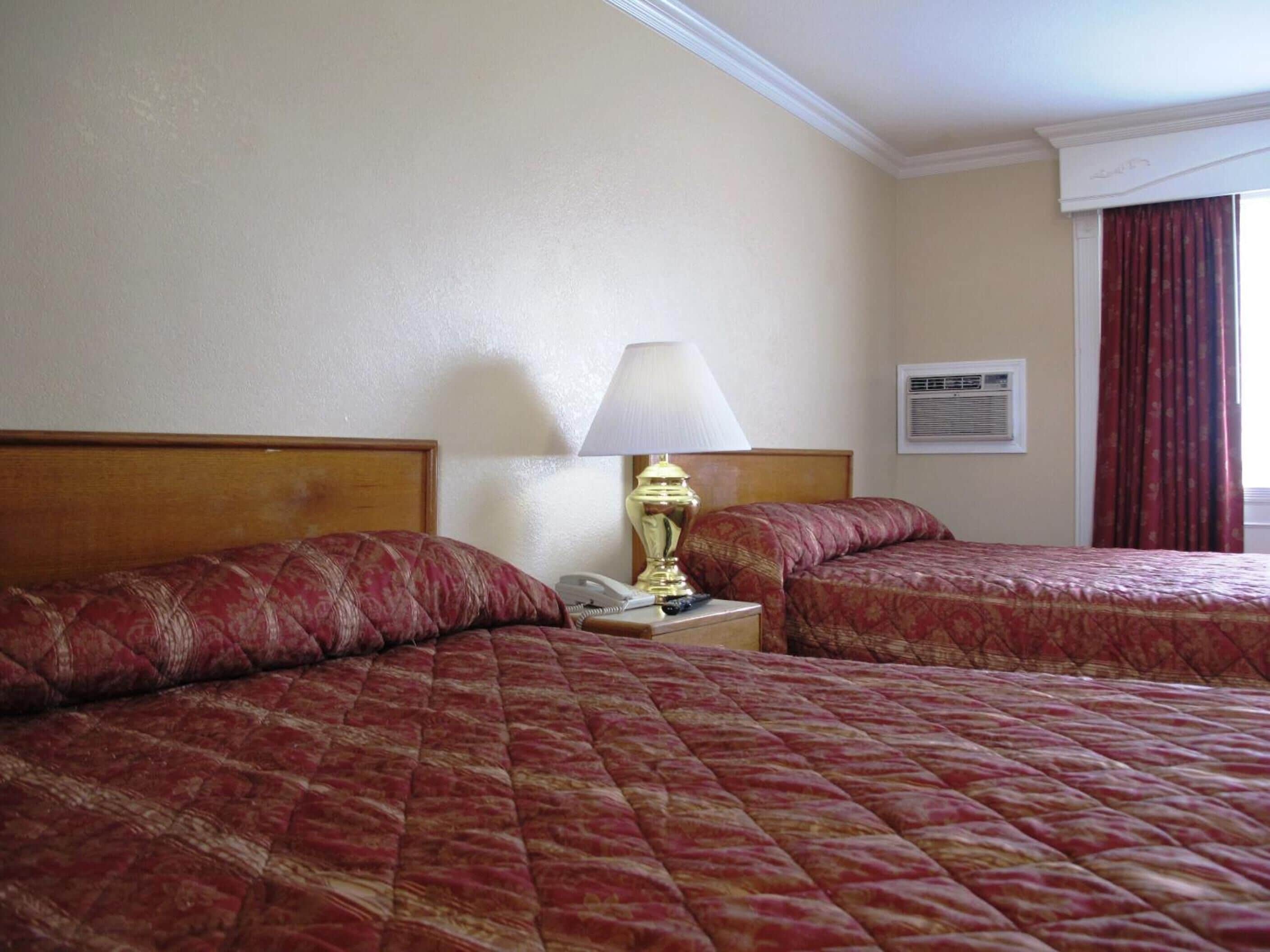 A hotel room with two beds, a nightstand with a lamp, a telephone, and an air conditioning unit.