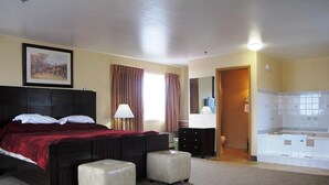 A hotel room with a large bed, two bedside tables, a nightstand, a lamp, a painting on the wall, a bathroom with a bathtub, and a shower area. - Merrill Field Inn (Anchorage)
