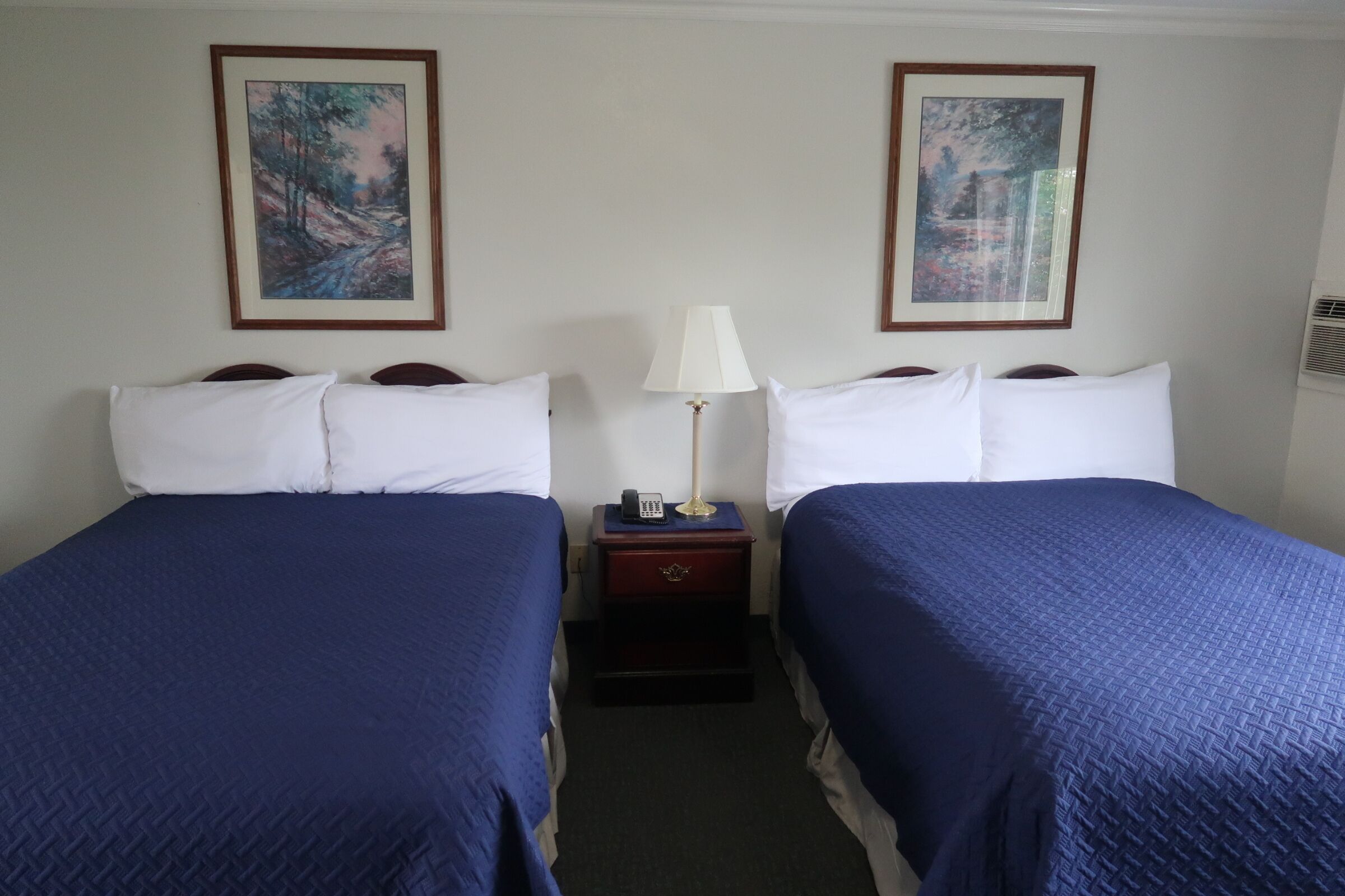 Een hotelkamer met twee bedden, elk voorzien van witte kussens en blauw bedlinnen. Daartussen staat een nachtkastje met een lamp en een telefoon.