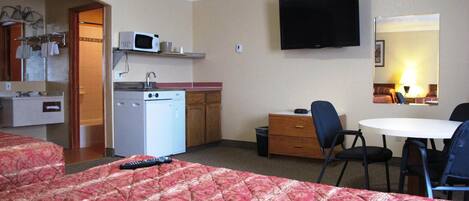 A hotel room with a bed, a desk, a chair, a TV, a microwave, a sink, and a small kitchenette.