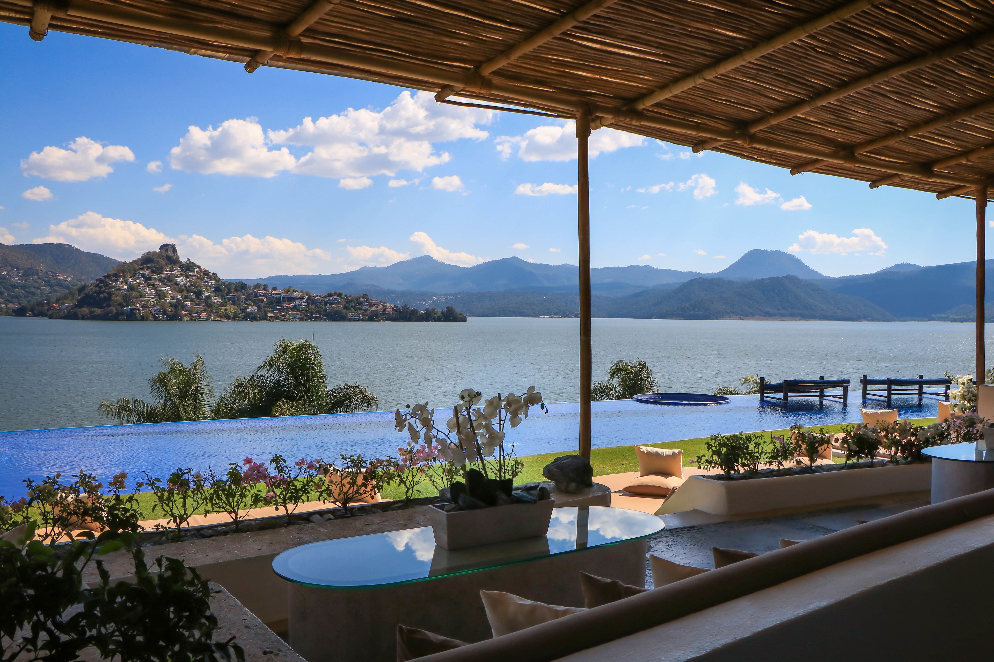 Una terraza con vistas a un lago y montañas, con mobiliario exterior y una mesa de vidrio.