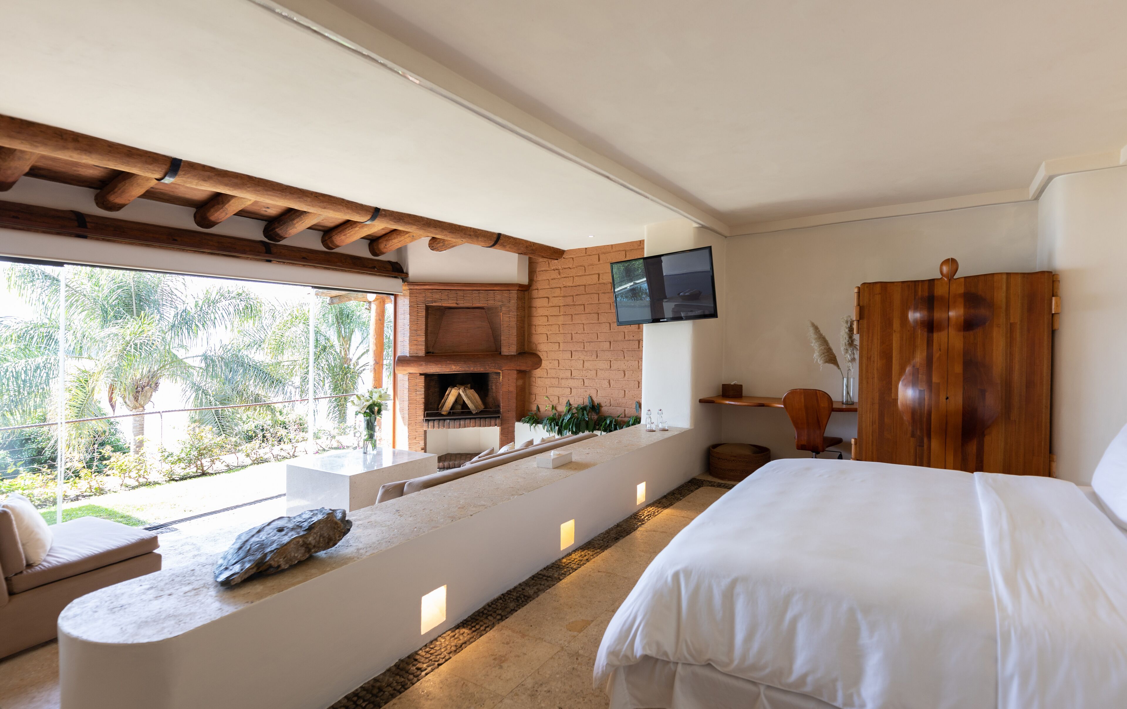 A bedroom with a bed, a TV, a wooden wardrobe, a brick fireplace, and a balcony with a view of palm trees.