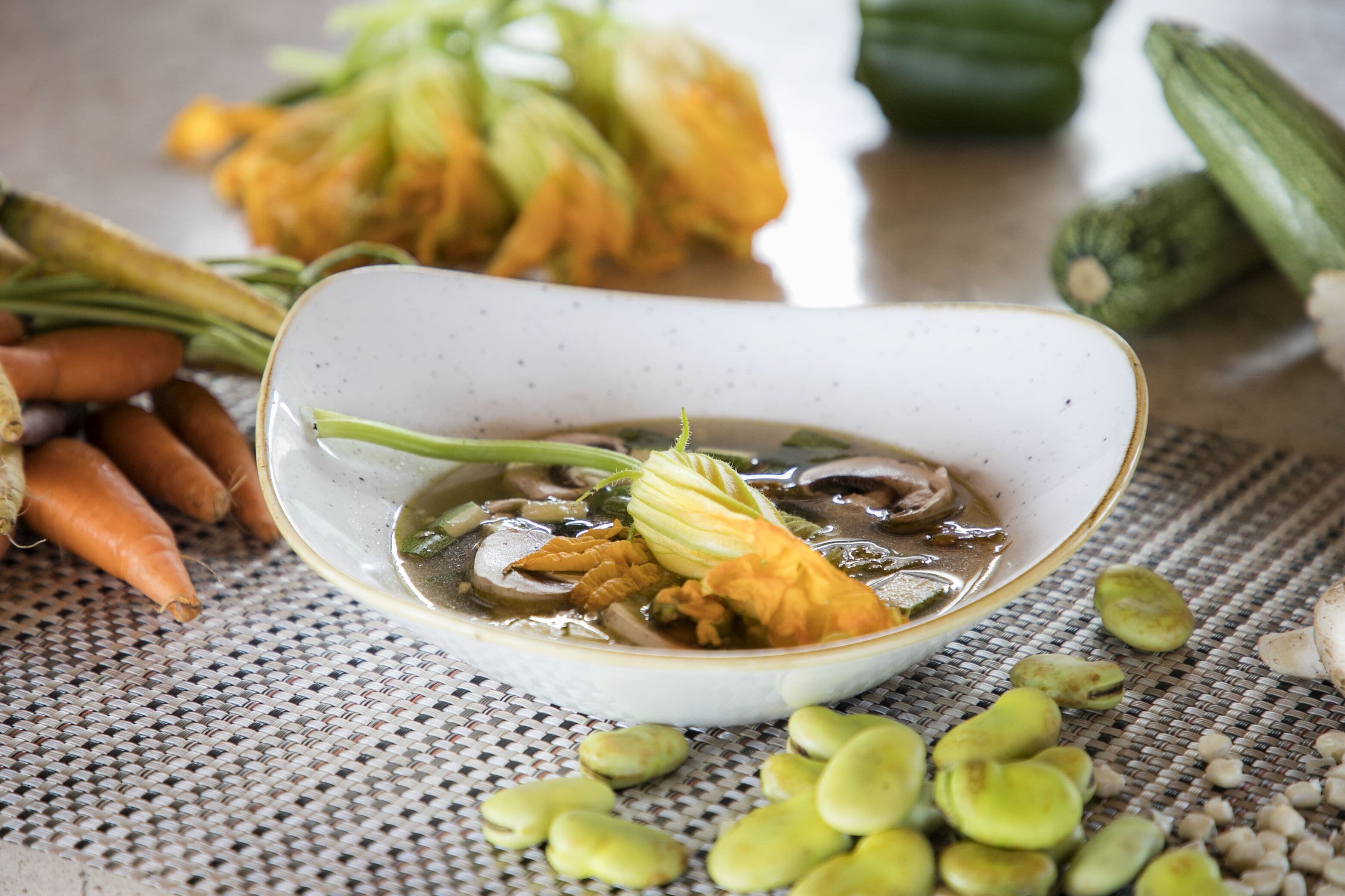 A bowl of soup with vegetables and herbs on a woven mat.