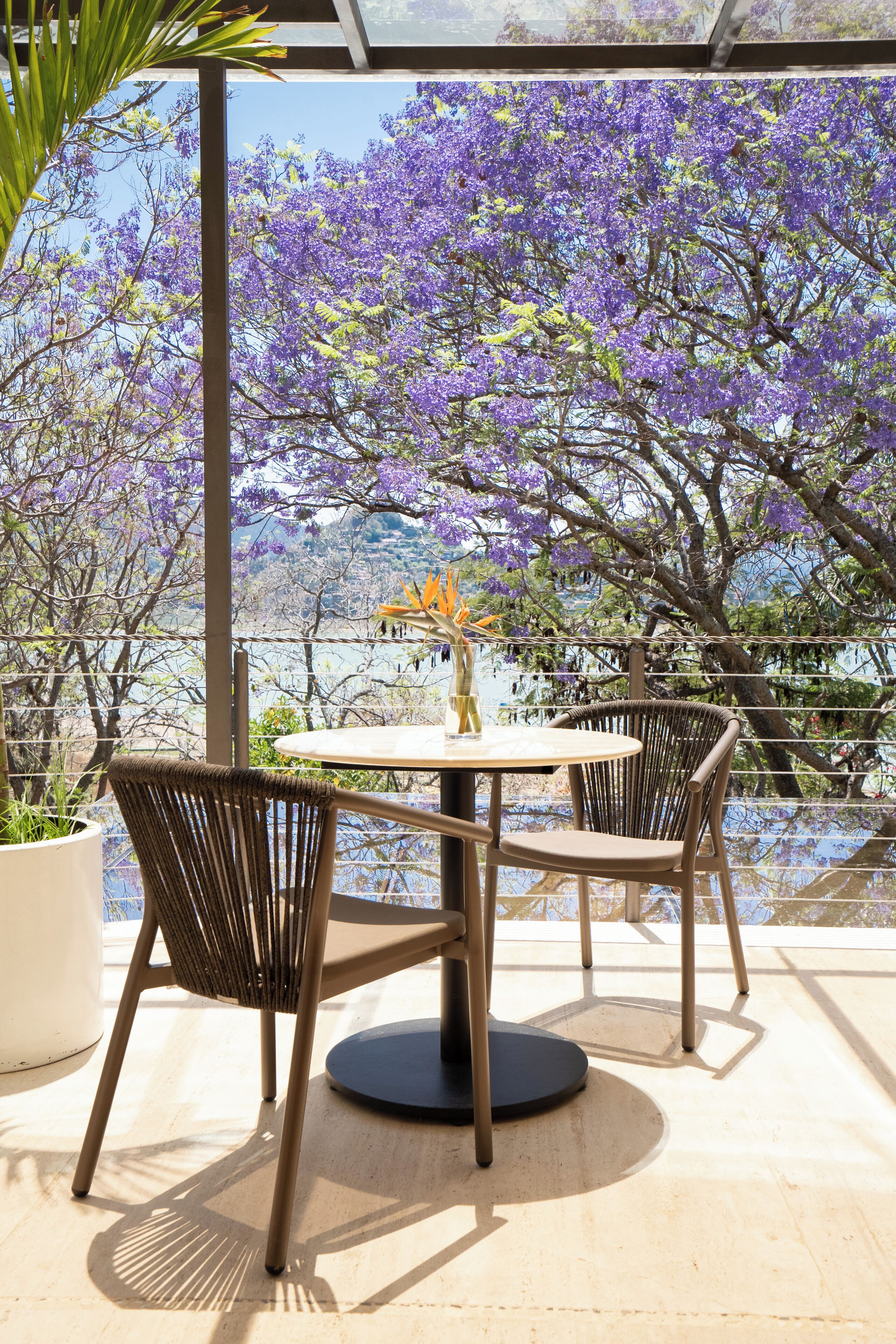 Un área de estar al aire libre con una mesa redonda y dos sillas, rodeada por un recinto de vidrio que ofrece vistas a árboles de flores moradas y un cuerpo de agua.