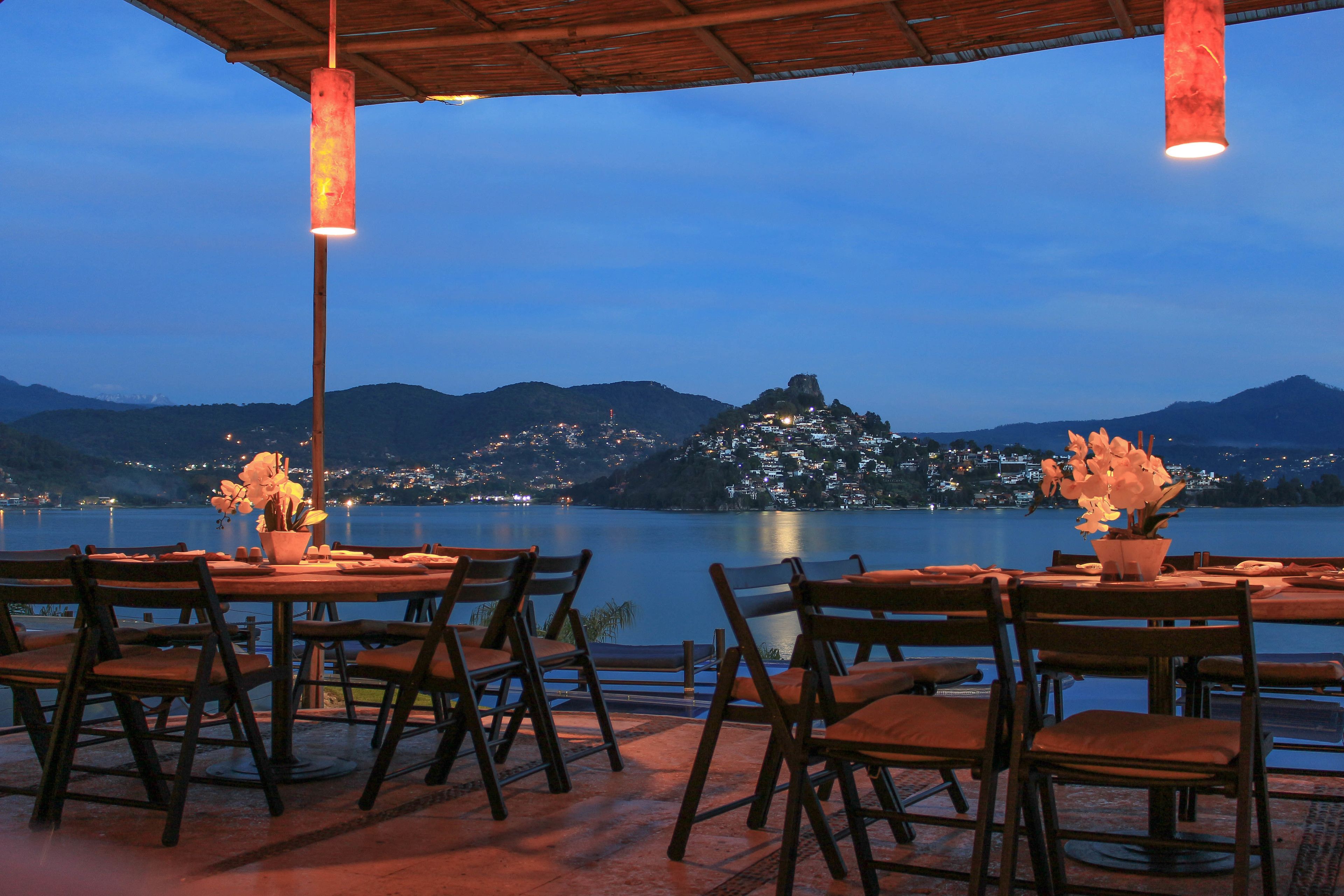 A terrace with tables and chairs overlooking a lake and a hill with buildings.