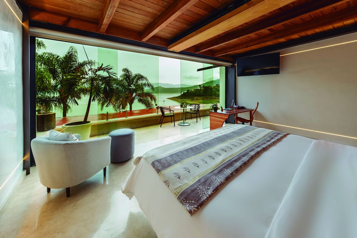 Ein modernes Schlafzimmer mit einem großen Bett, Blick auf Palmen und ein Gewässer durch raumhohe Fenster, einer kleinen Sitzecke und einem Esstisch.
