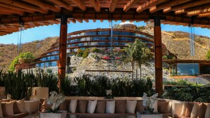 A modern outdoor seating area with a glass table and sofa, surrounded by lush plants and a view of a multi-story building with a dome on top. - El Santuario Resort & Spa (Valle de Bravo)