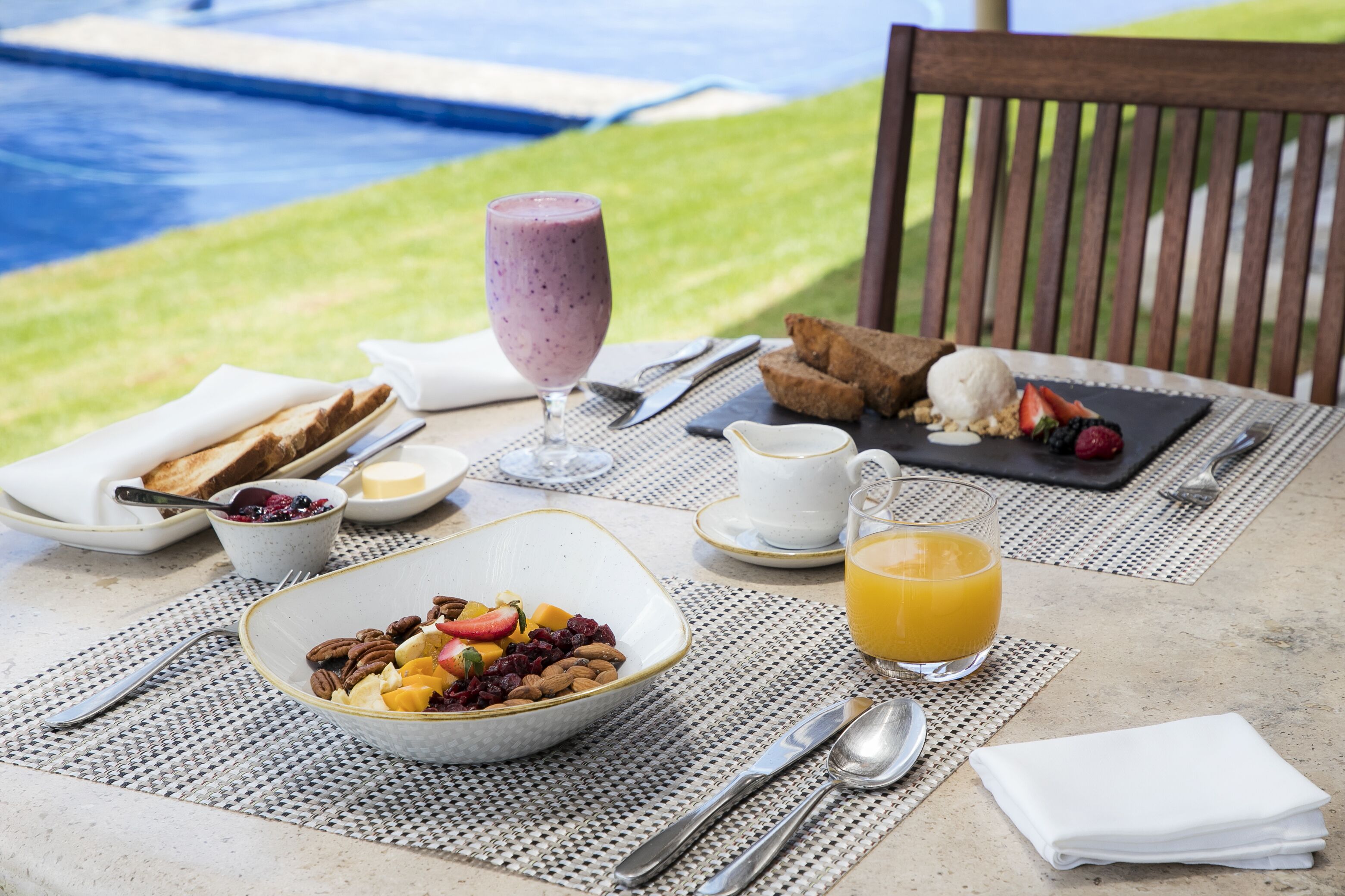 A breakfast setting with a pool view, including a bowl of cereal, toast, fruit, a glass of orange juice, and a glass of smoothie.