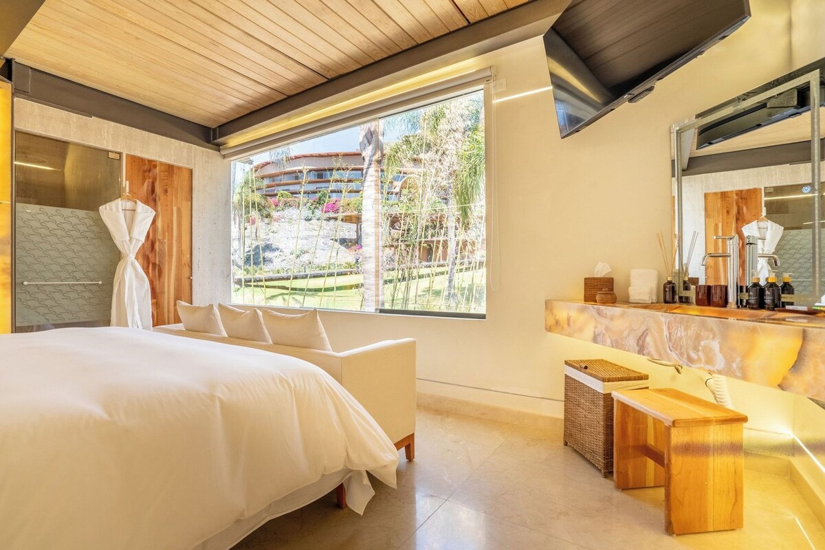 A modern hotel room with a large bed, a sitting area, a bathroom with a vanity, and a view of a garden.